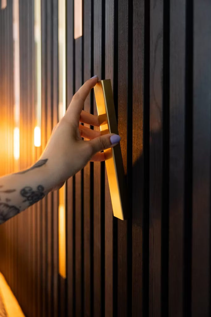 Wytatuowana dłoń z jasnofioletowymi paznokciami chwyta złoty uchwyt na eleganckiej, ciemnej ścianie z akustycznych paneli drewnianych Akupanel, podkreślonej ciepłym oświetleniem.
